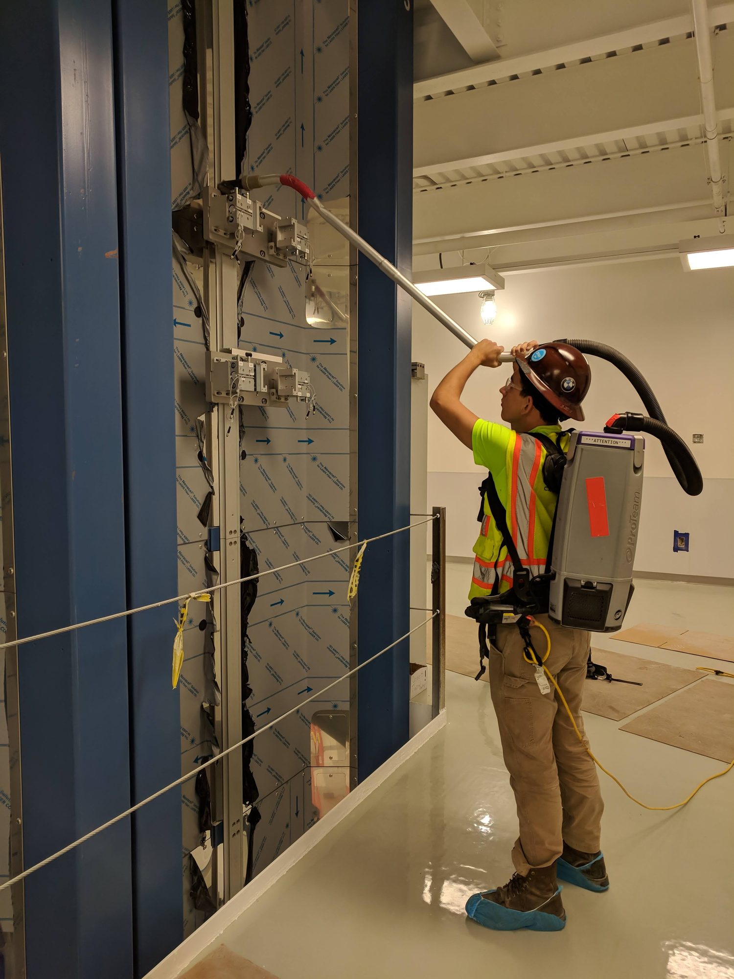A CMI tech in harness uses a backpack vacuum and extension wand to dust high steel on a new fiber draw tower.