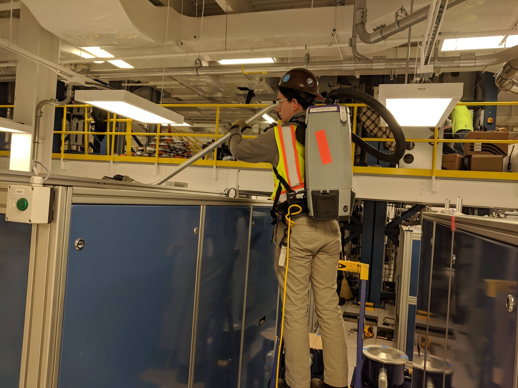 A CMI tech uses a backpack vac wand to clean the tops of new equipment enclosures under a mezzanine.