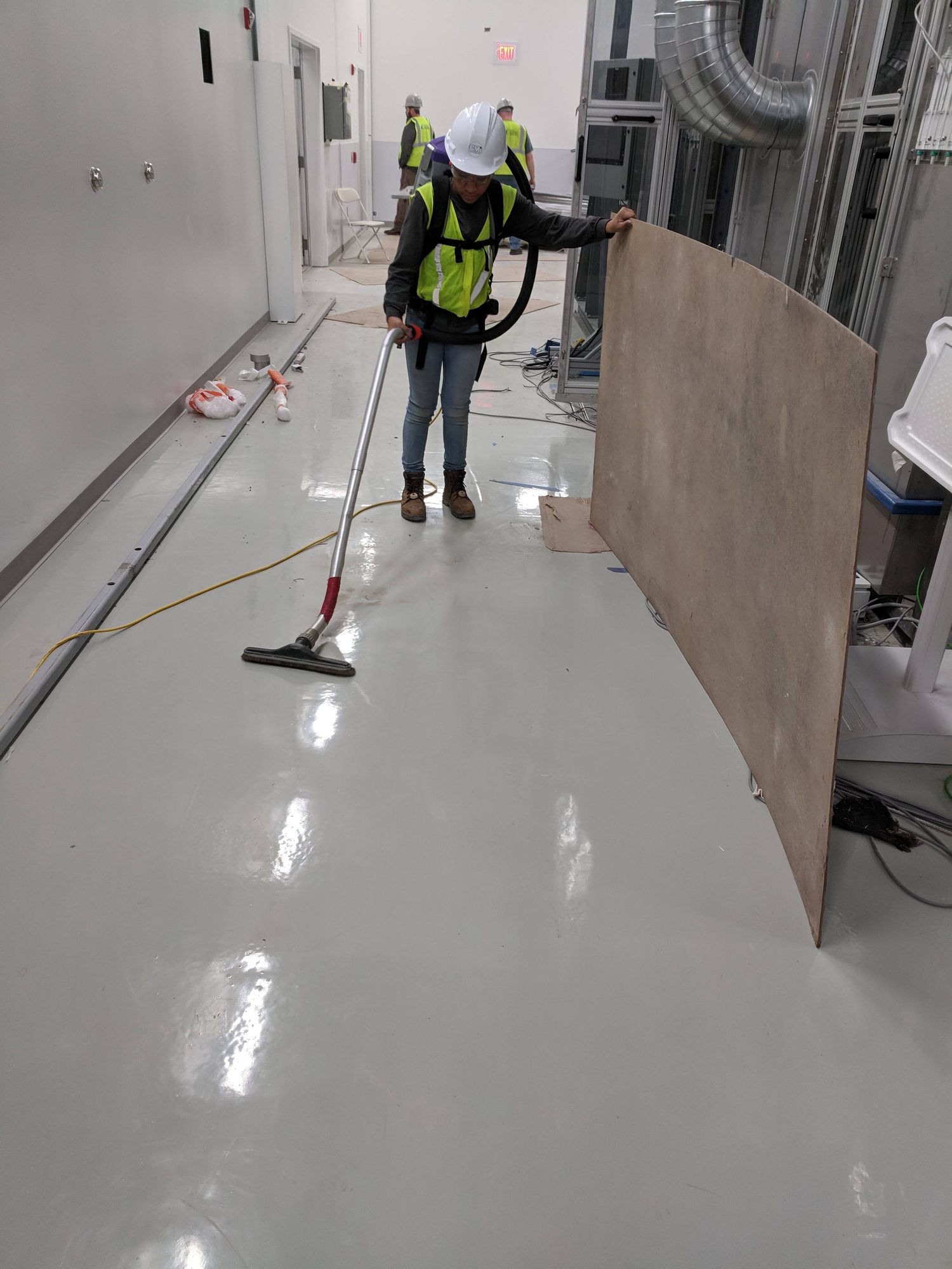 A CMI tech in hard hat and vest vacuums a freshly coated corridor floor with a backpack vac during final clean of a new fiber plant.