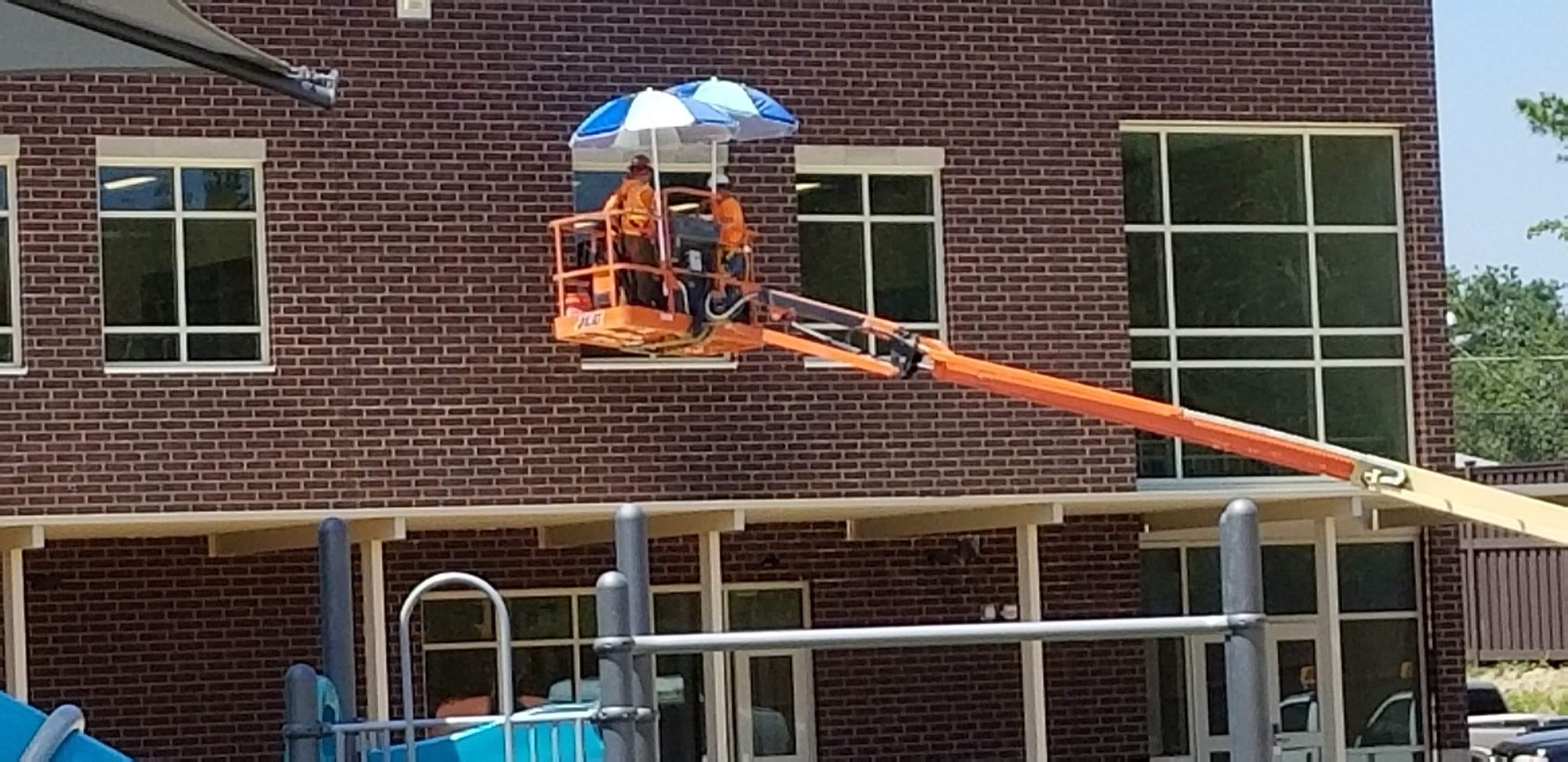 Two CMI techs in a boom lift basket, shaded by an umbrella, wash second-floor windows on a new school.