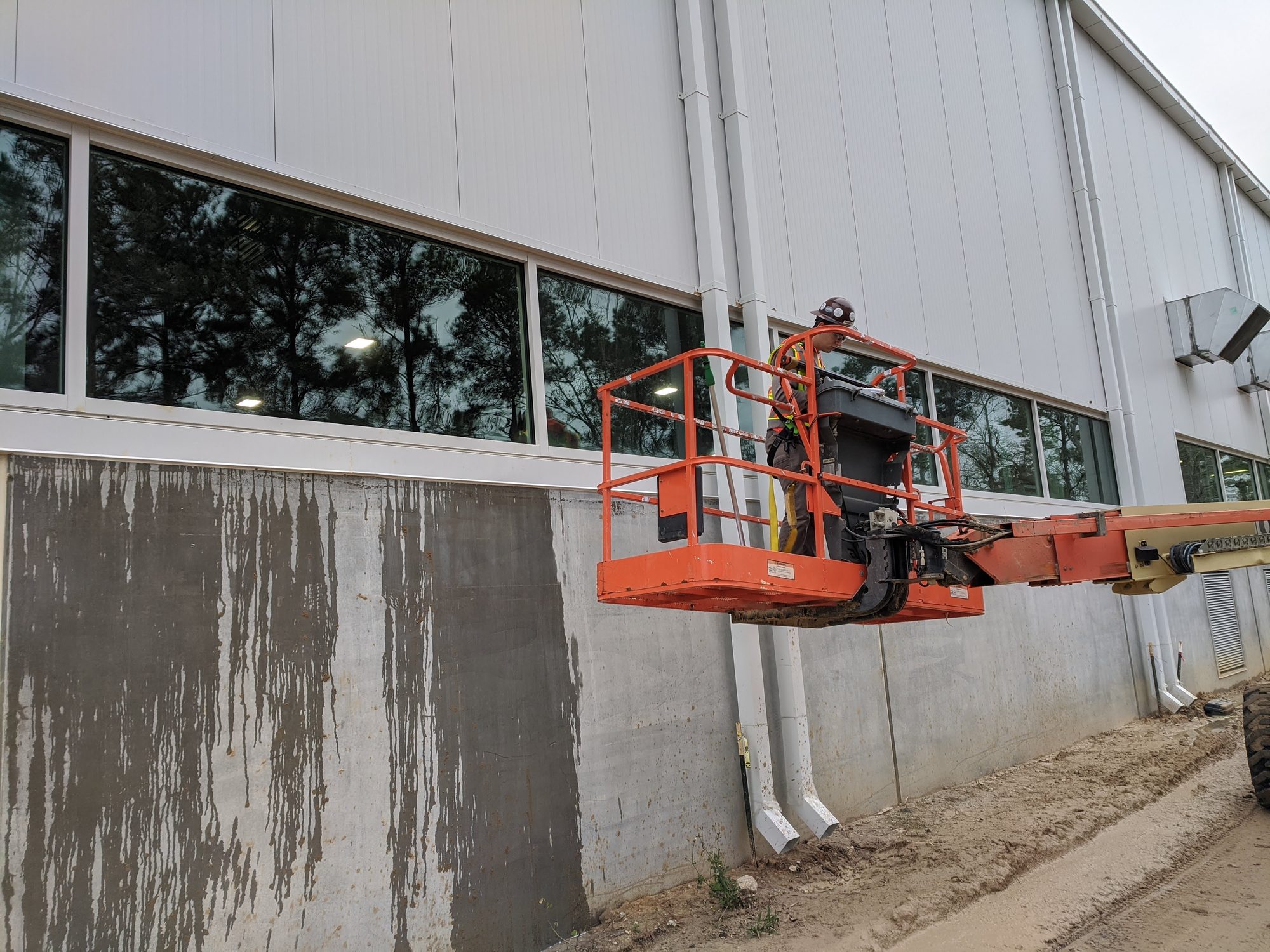 A CMI tech on a boom lift washes construction staining off a new plant's concrete wall, with clean and stained sections side by side.