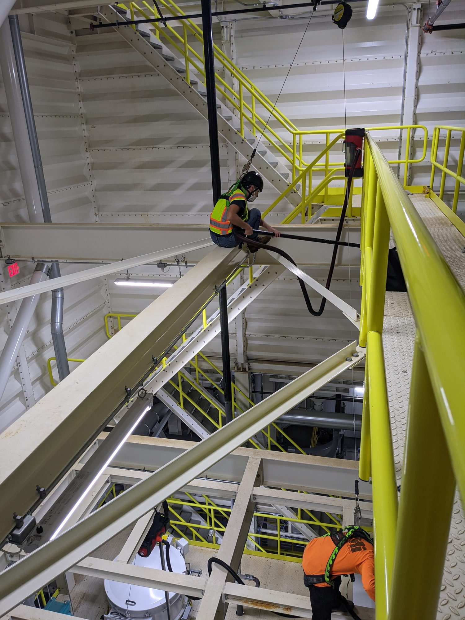 Harnessed CMI techs vacuum construction dust off steel framing and platforms inside a new production plant.