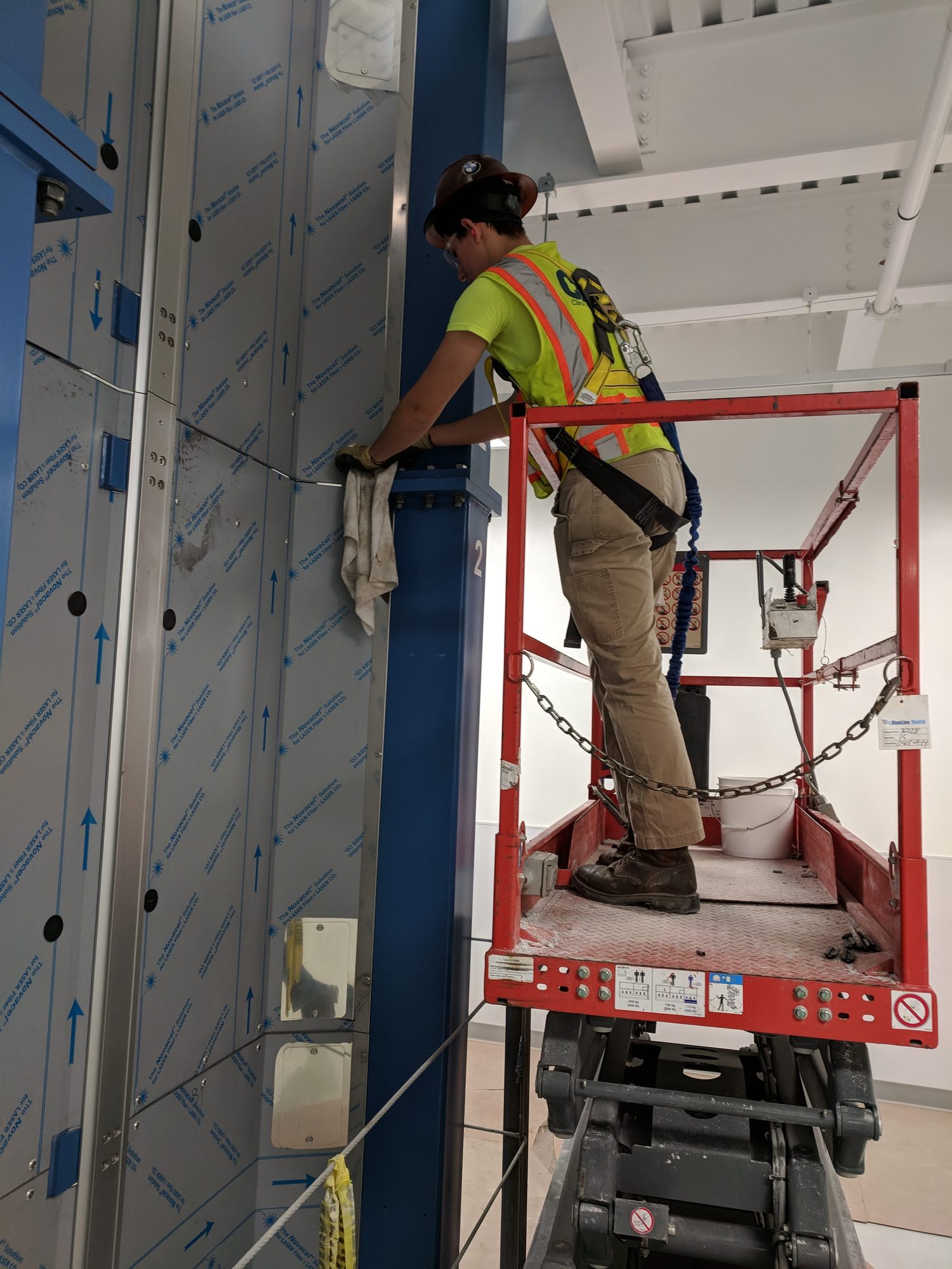 A harnessed CMI tech on a scissor lift wipes down a steel column on a new fiber draw tower.