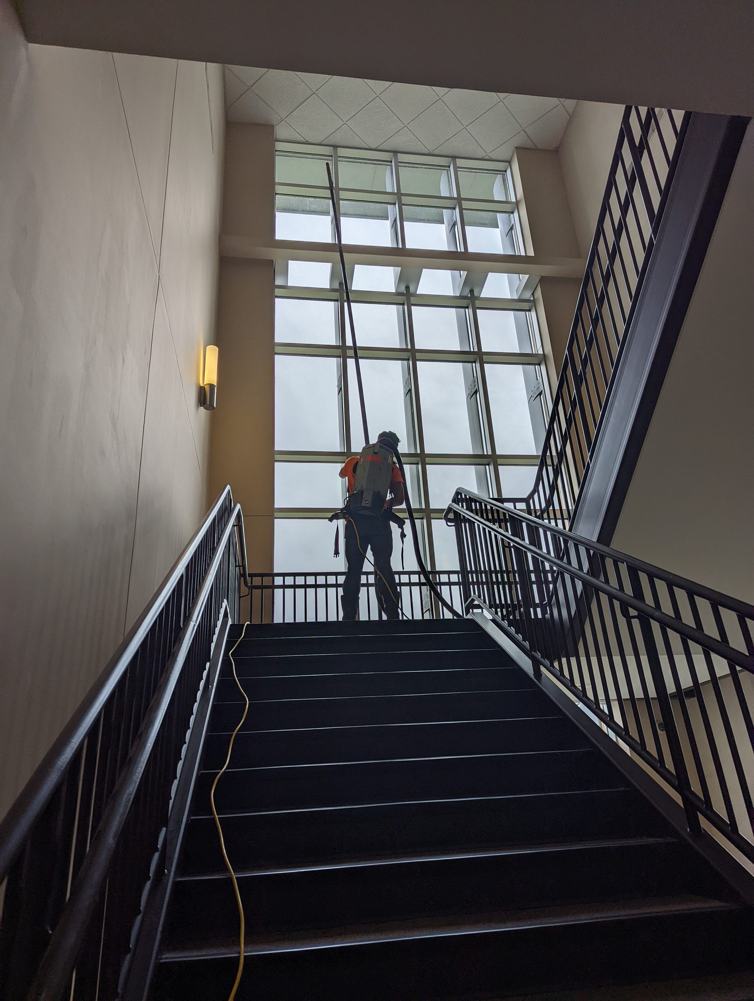 A CMI tech on a stair landing cleans a tall curtain-wall window with an extension pole during final clean.