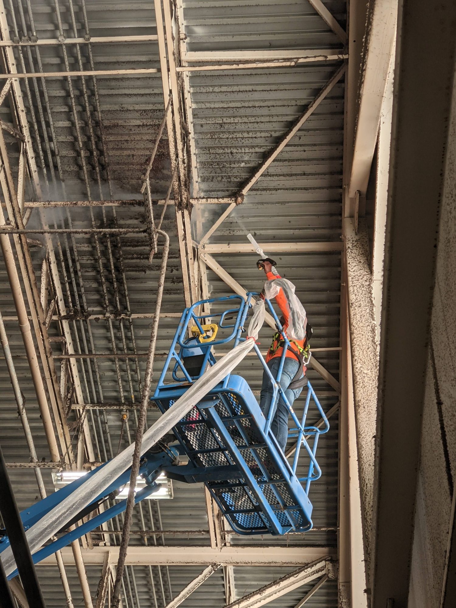 Harnessed tech in a boom lift dry ice blasts lint-caked roof trusses and deck high above a textile plant floor.