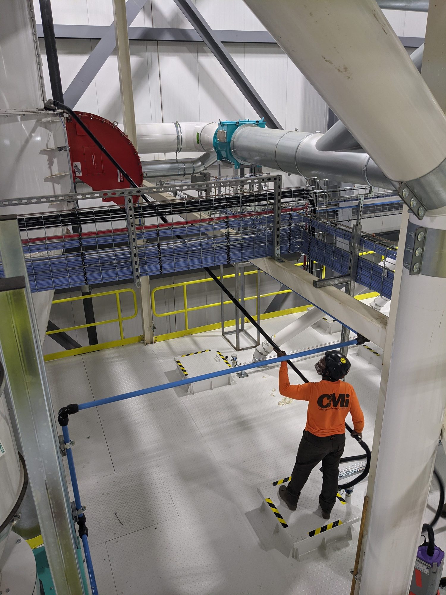 CMI tech pole-vacuums overhead pipe racks and cable trays from a platform below.