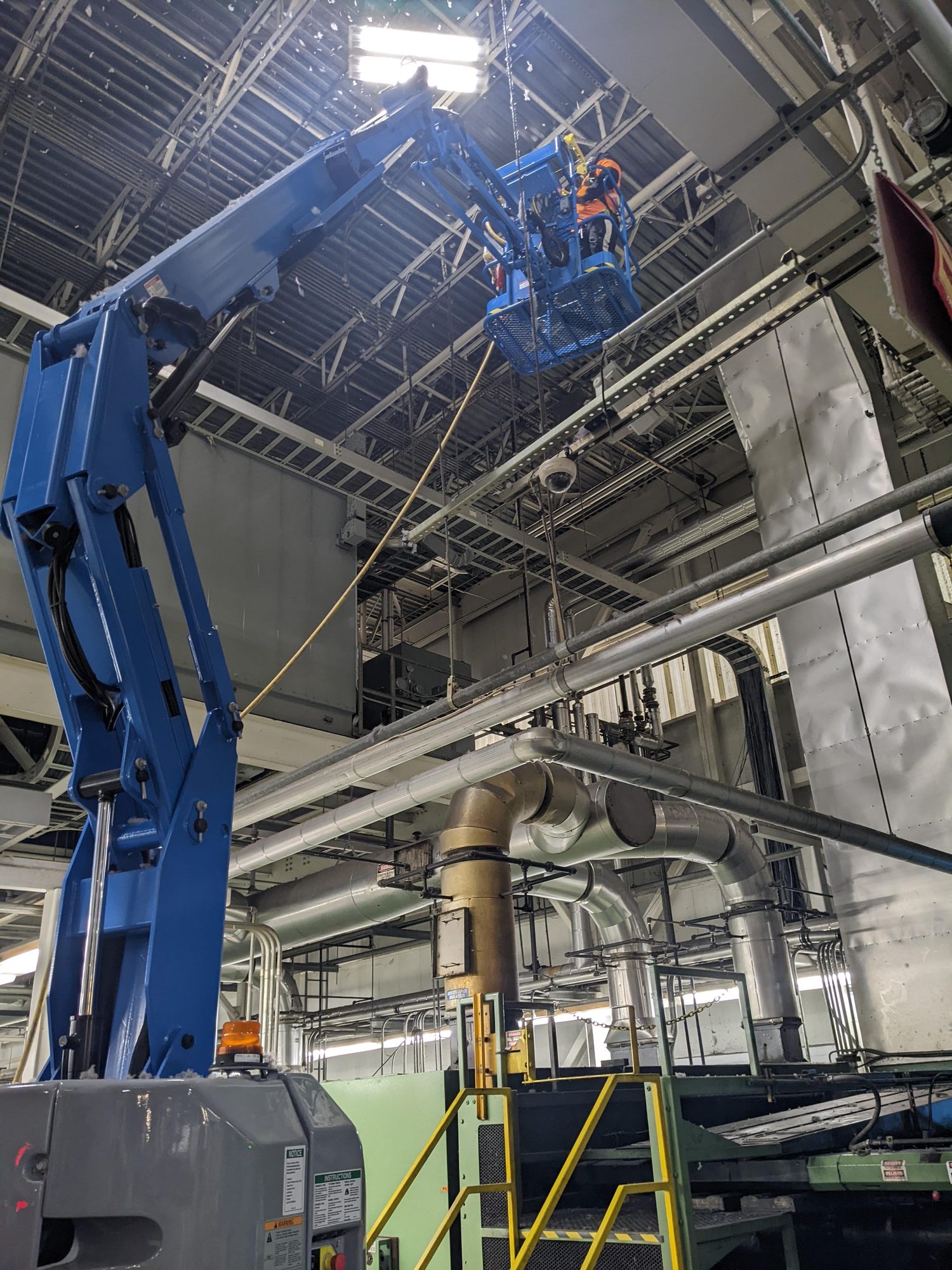A harnessed tech works from a boom lift high in a textile plant, cleaning lint off overhead ducts and steel.