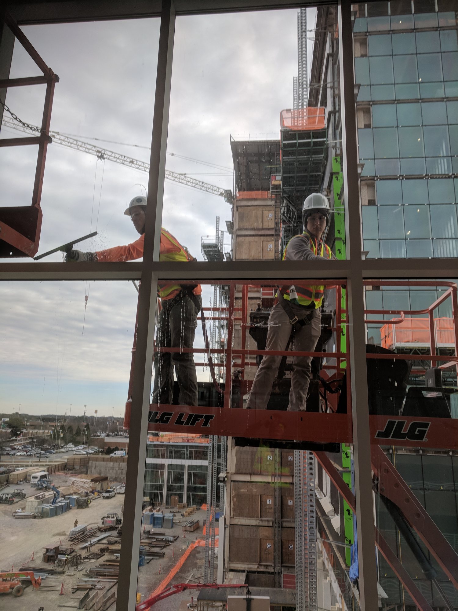 Two hard-hatted techs in a lift basket squeegee new curtain wall glass on a hospital tower under construction.
