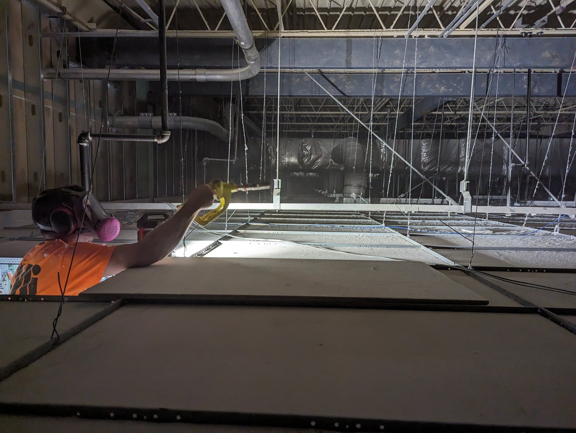 A CMI tech in a respirator works inside the ceiling plenum above a textile plant, lighting the lint-covered tiles before vacuuming.