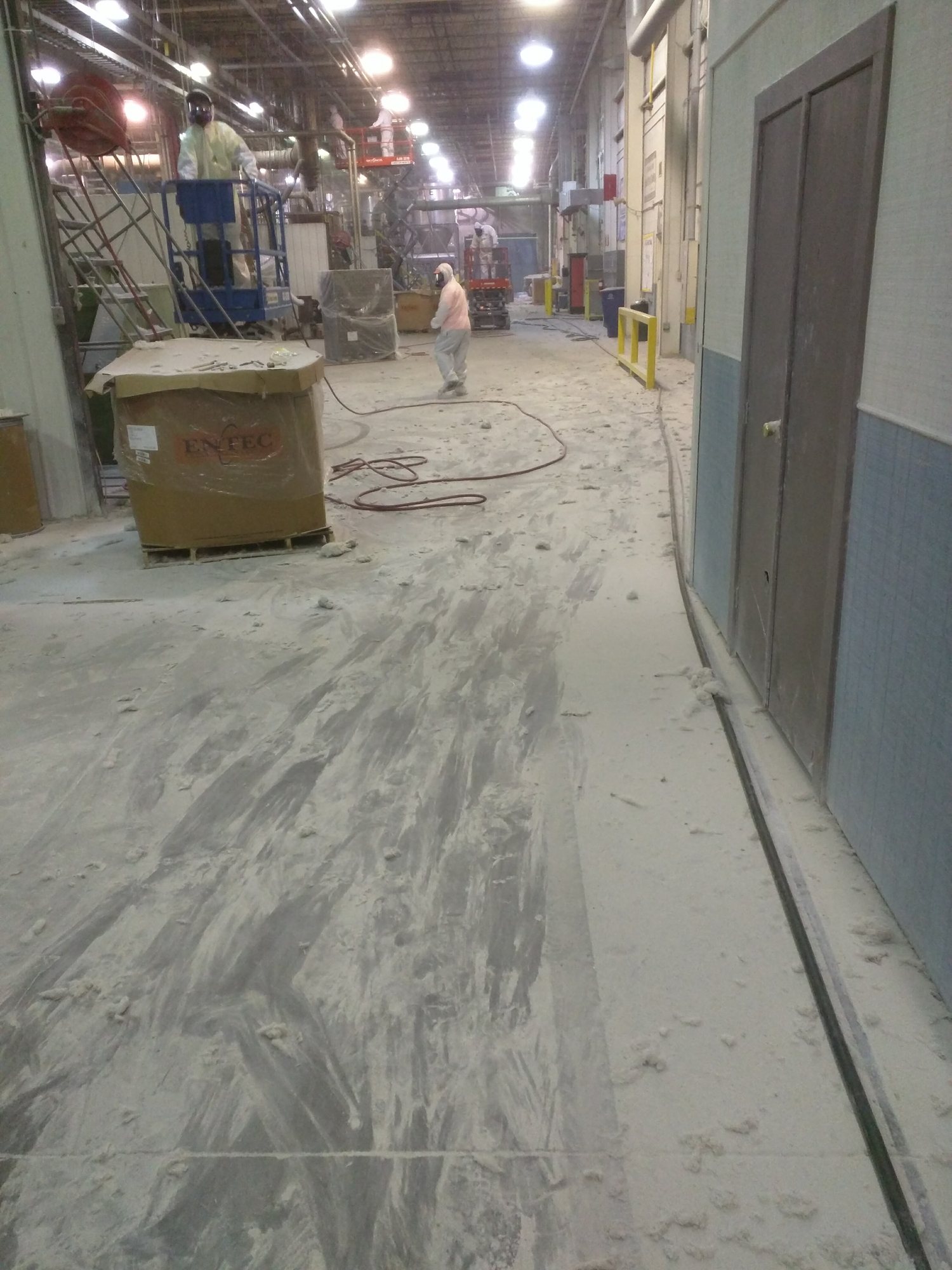 During an overhead lint cleanout, the aisle floor is covered in fallen fiber while crews in Tyvek work from lifts.