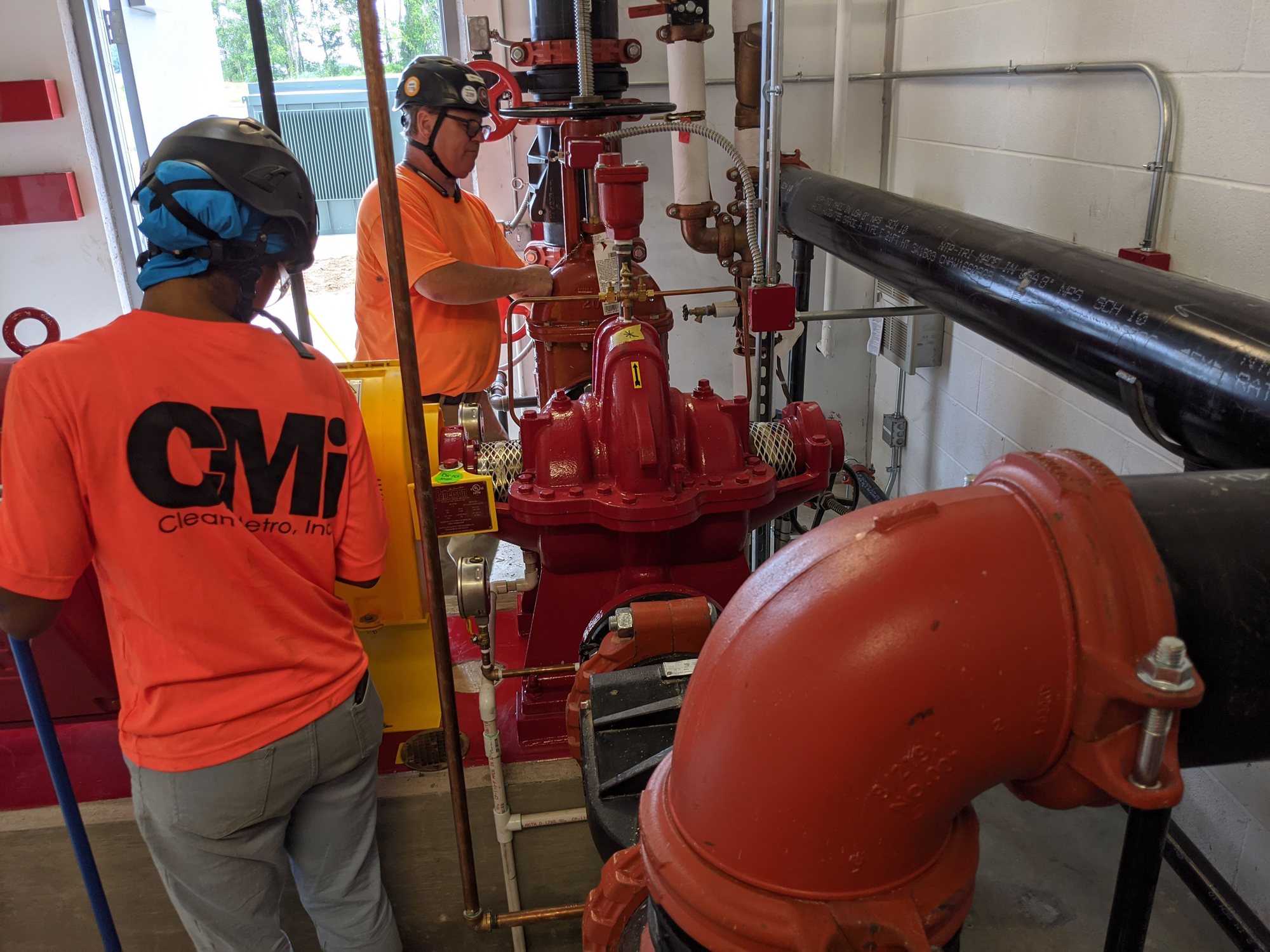 Two CMI techs in hard hats clean the fire pump and piping in a new pump room before turnover.