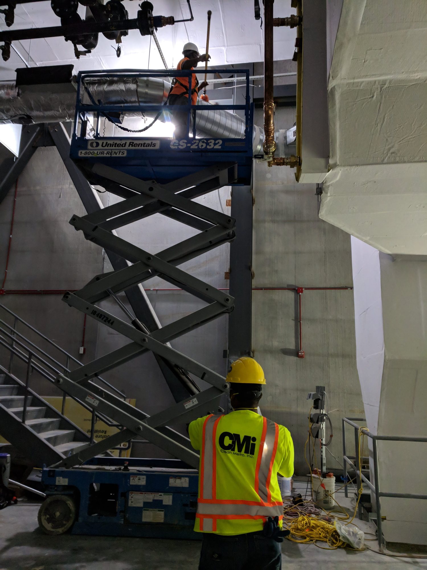 A CMI spotter in a branded vest watches a teammate clean overhead piping from a scissor lift.