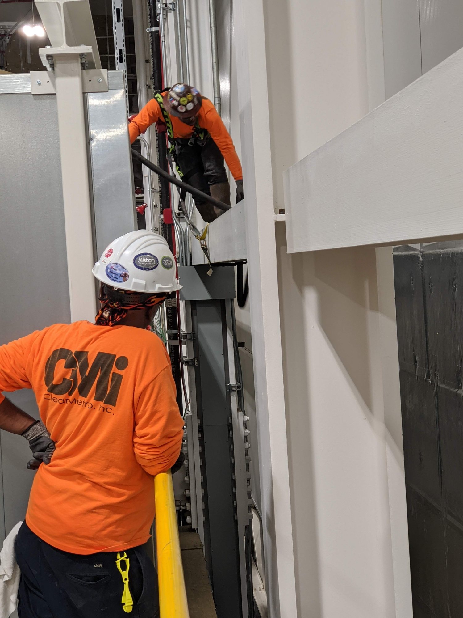 CMI spotter watches a harnessed coworker work down a narrow mechanical shaft.