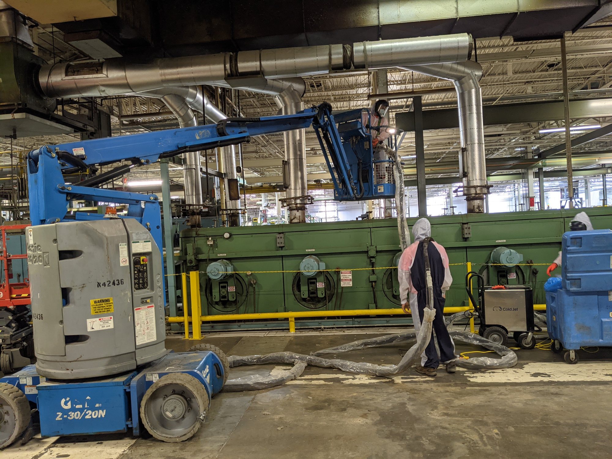 Tech in a boom lift blasts overhead ductwork while a ground tech tends the blast hose beside the textile range.