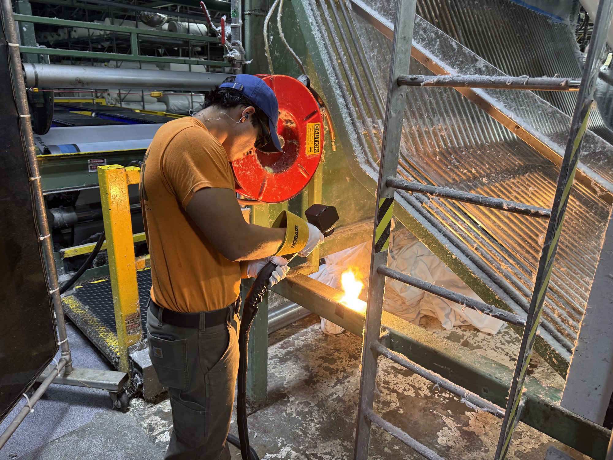 A CMI tech laser cleans a lint-caked steel crossmember in a textile plant, the bright flash marking the cleaning point.