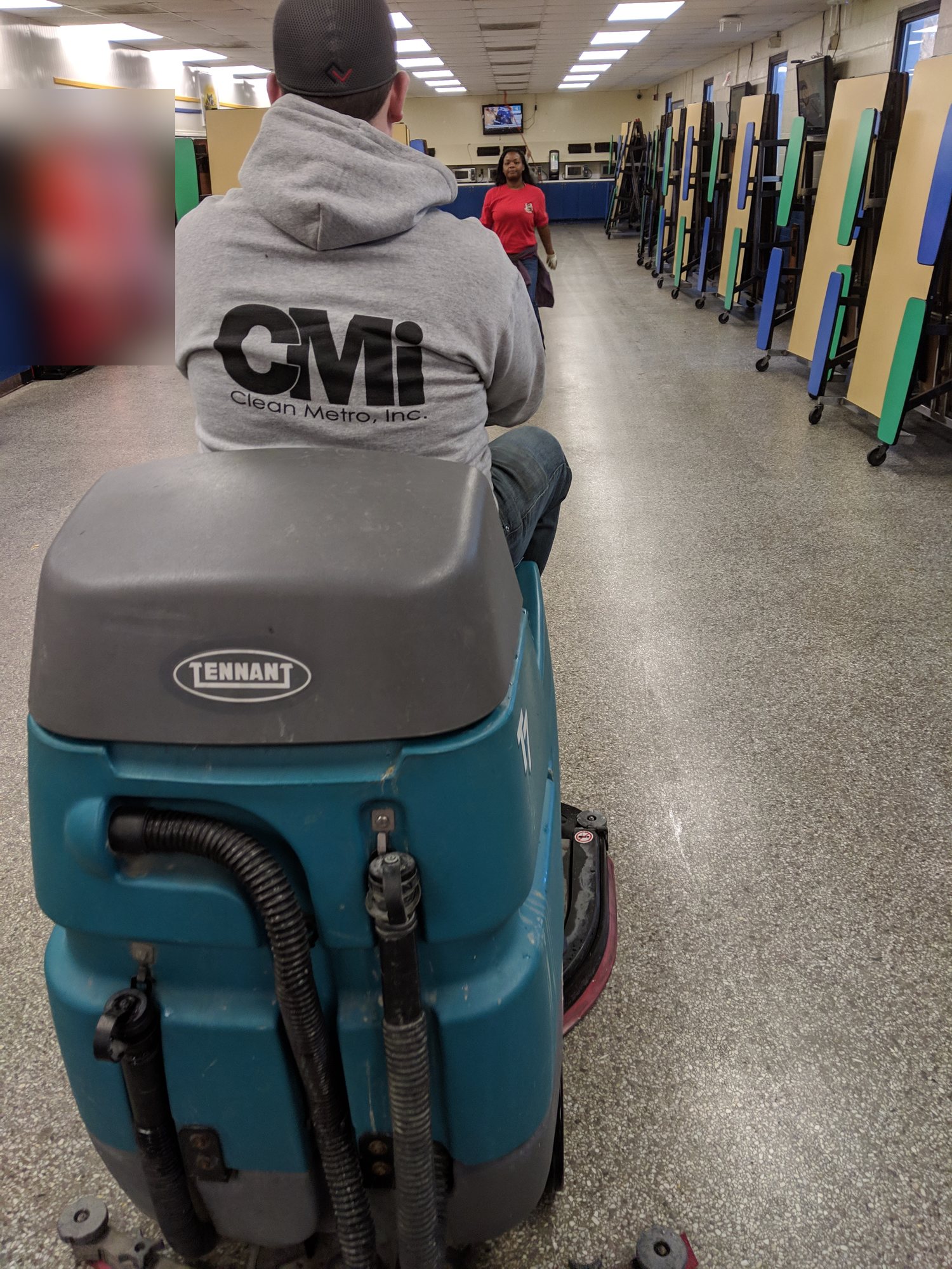 CMI tech runs a ride-on scrubber down a long cafeteria floor.