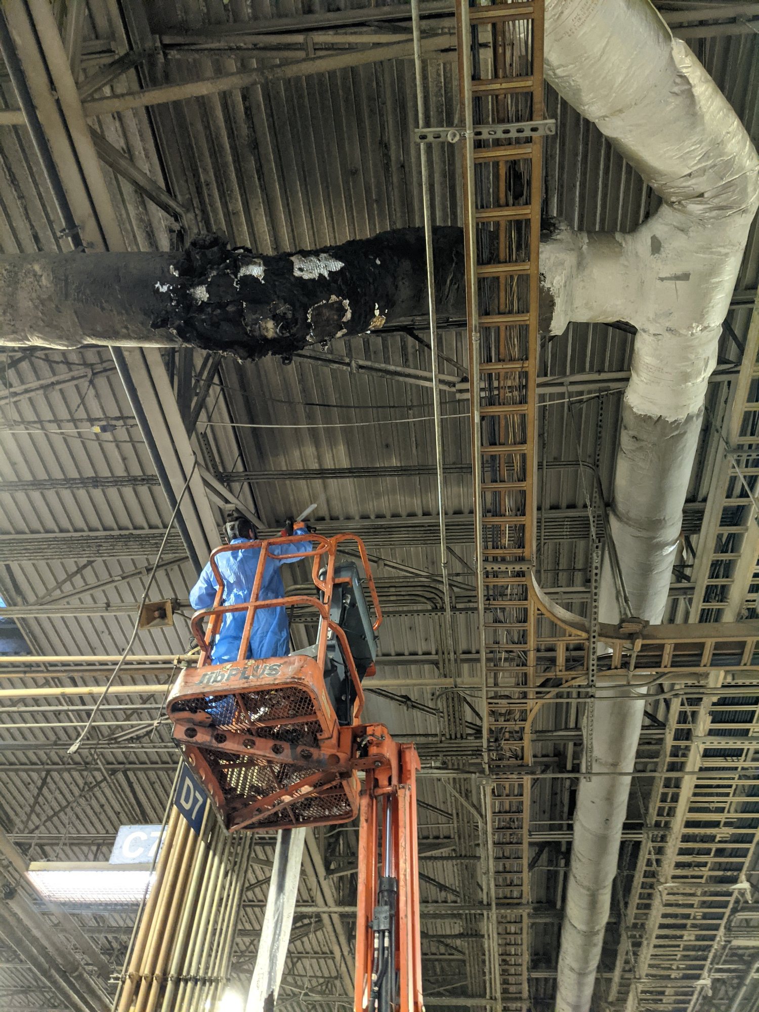 A CMI tech works from a lift beside charred pipe insulation after a fire in a textile plant, before damaged material removal.