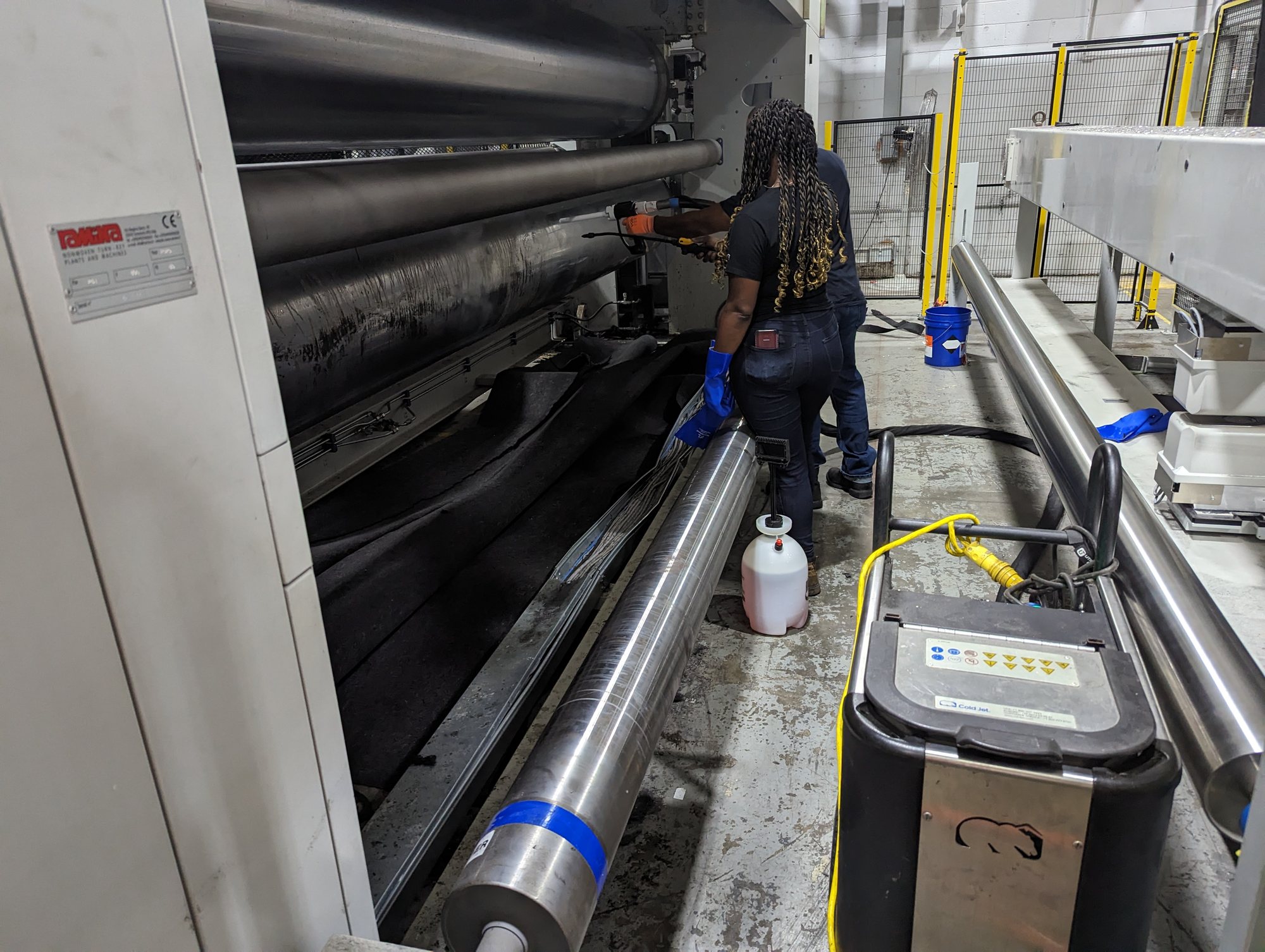 Two CMI techs in gloves clean residue off textile machine rollers with a pump sprayer and wet vac.