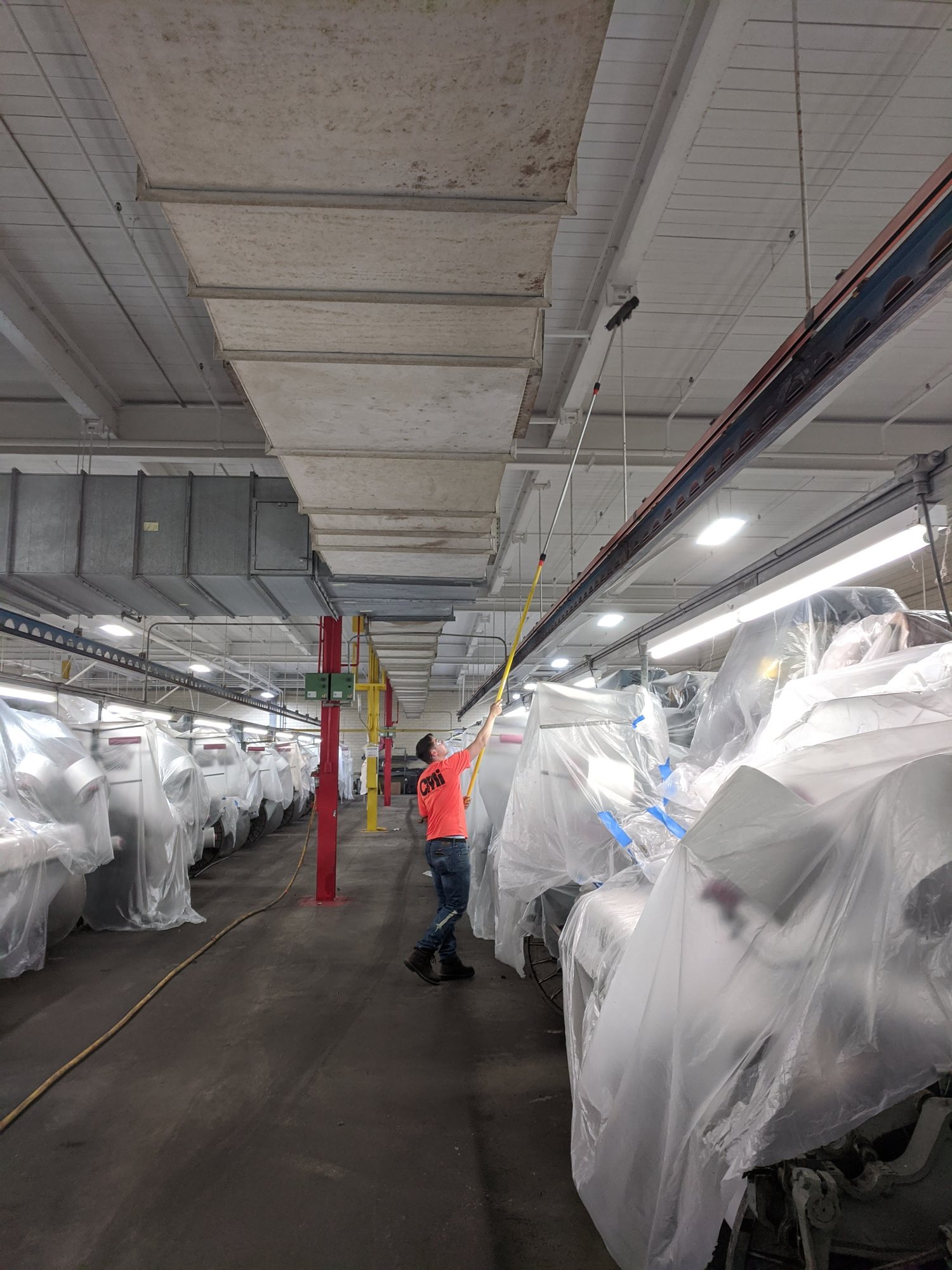 CMI tech dusts overhead ductwork with an extension pole above rows of plastic-wrapped machines.