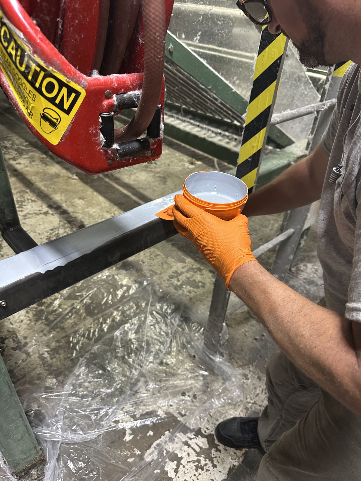 A tech brushes primer onto the laser-cleaned beam, with the floor protected by plastic sheeting.