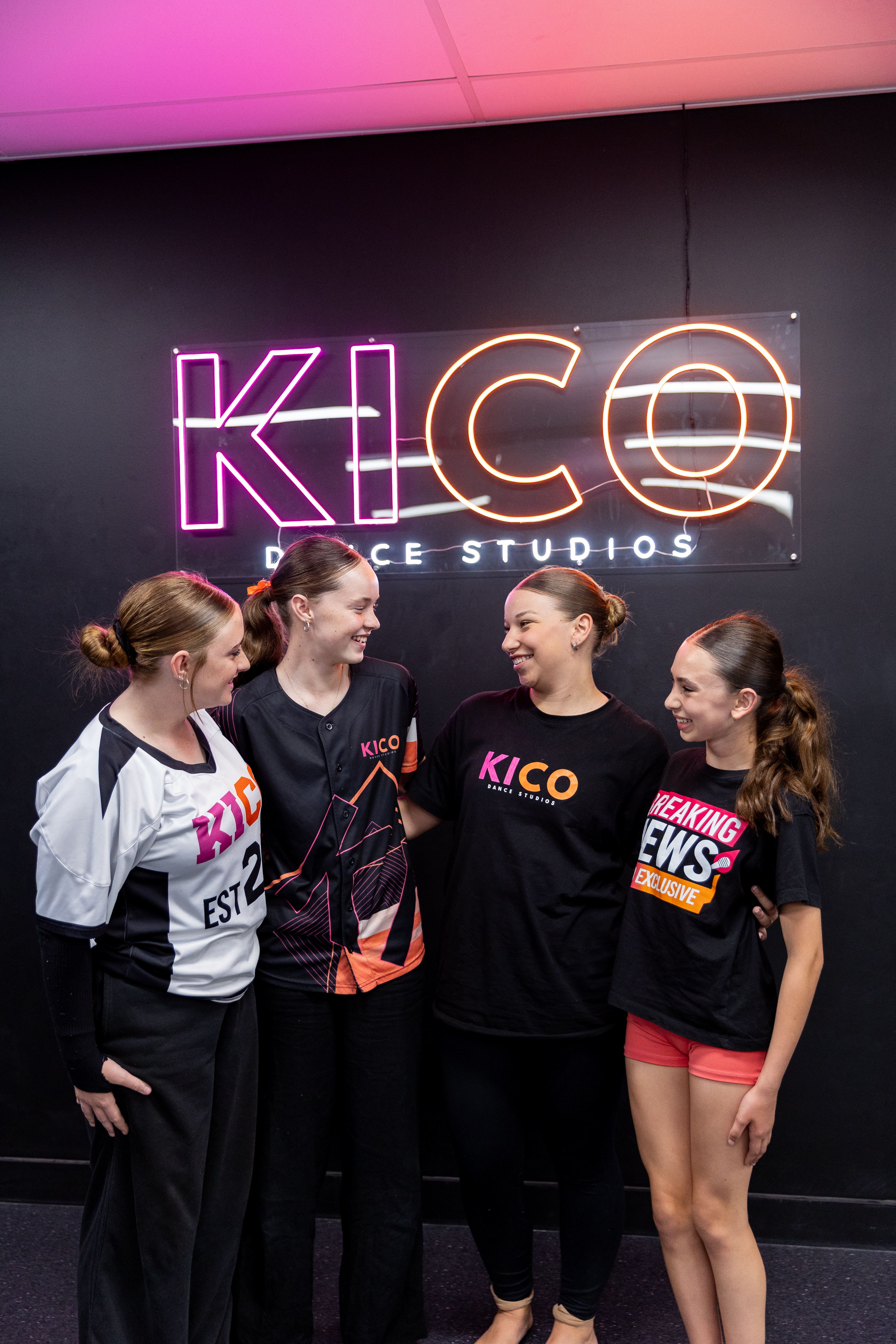 "Smiling KICO dancers showing teamwork in front of the neon studio sign." "Smiling KICO dancers showing teamwork in front of the neon studio sign."