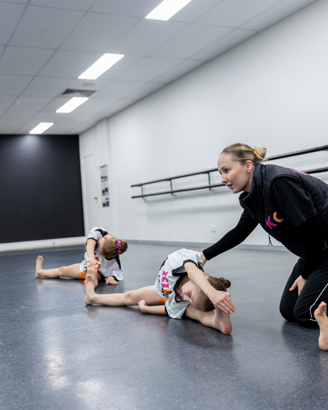 Dance teacher encouraging a young student performing a side stretch in a straddle split during a studio flexibility class.