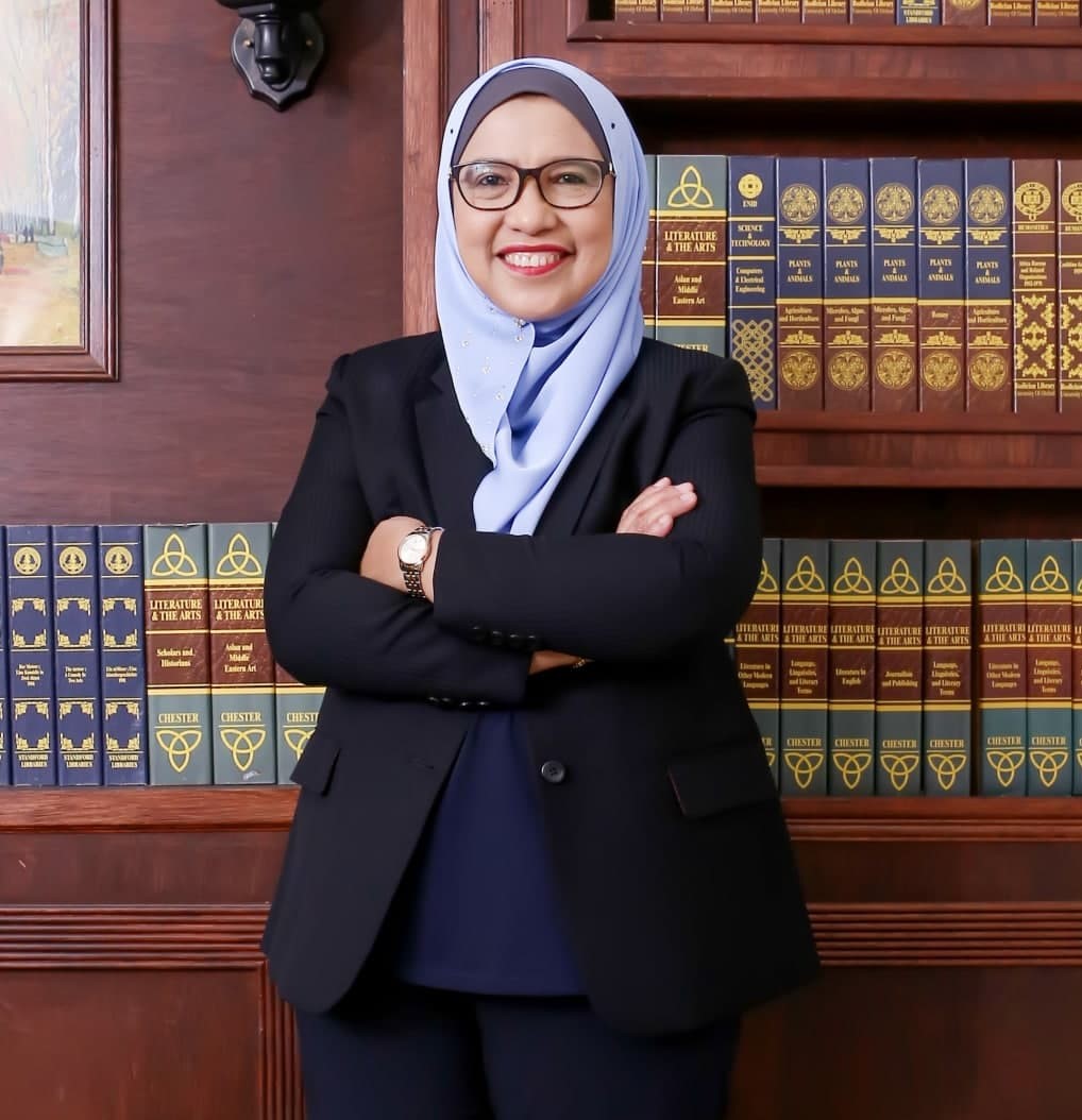 A'tikah Ahmad berpakaian blazer hitam dan tudung biru muda di hadapan rak buku