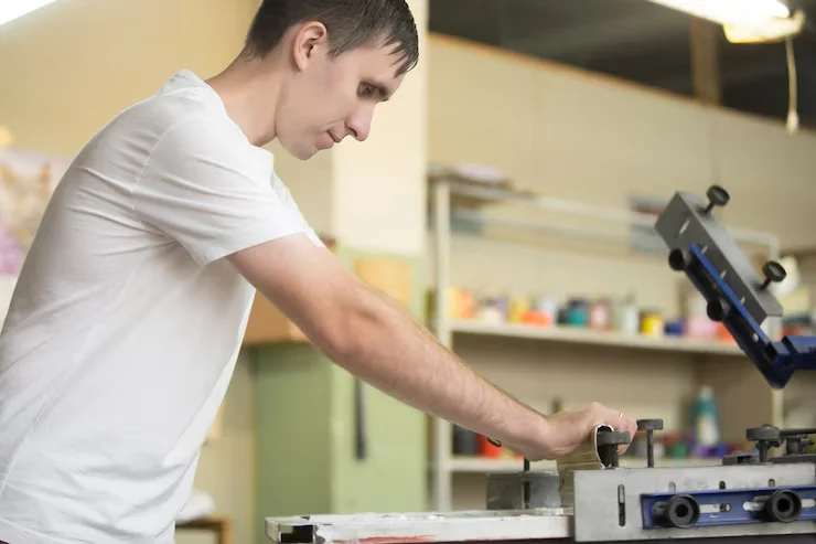 A man in a white t-shirt operating a screen printing machine, focused on the task in a brightly lit workshop.