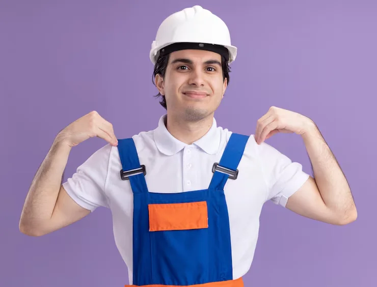 A young male builder in a white hard hat and blue overalls with orange accents, confidently pulling his suspenders, looking at the camera.