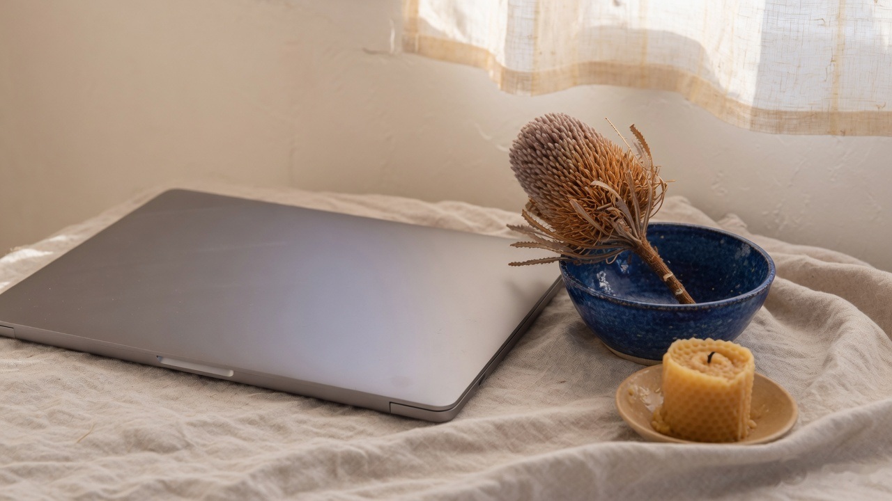 A closed laptop, a bowl of dried seed pods and a candle on a linen cloth by a window