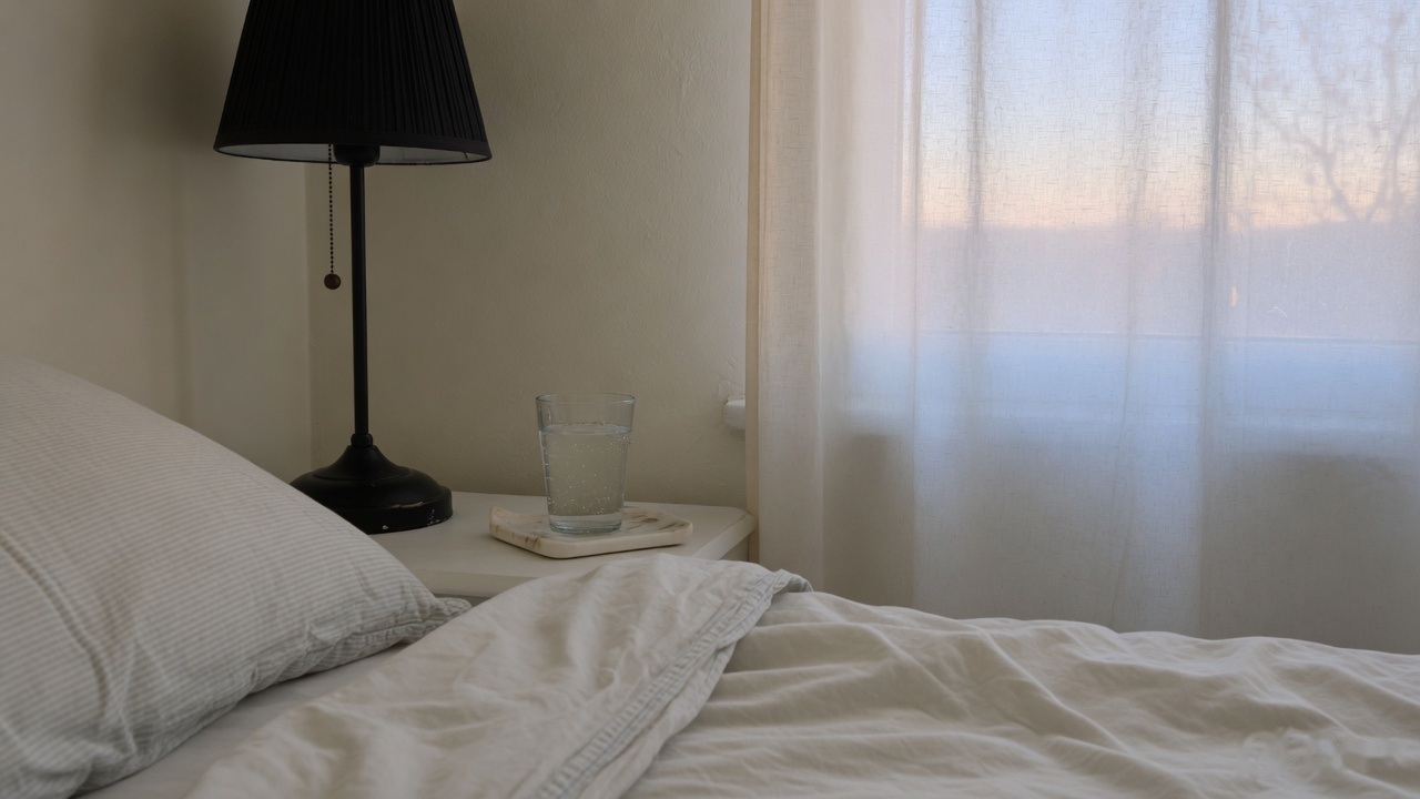 A bedside lamp and a glass of water beside a made bed, with morning light through the curtain