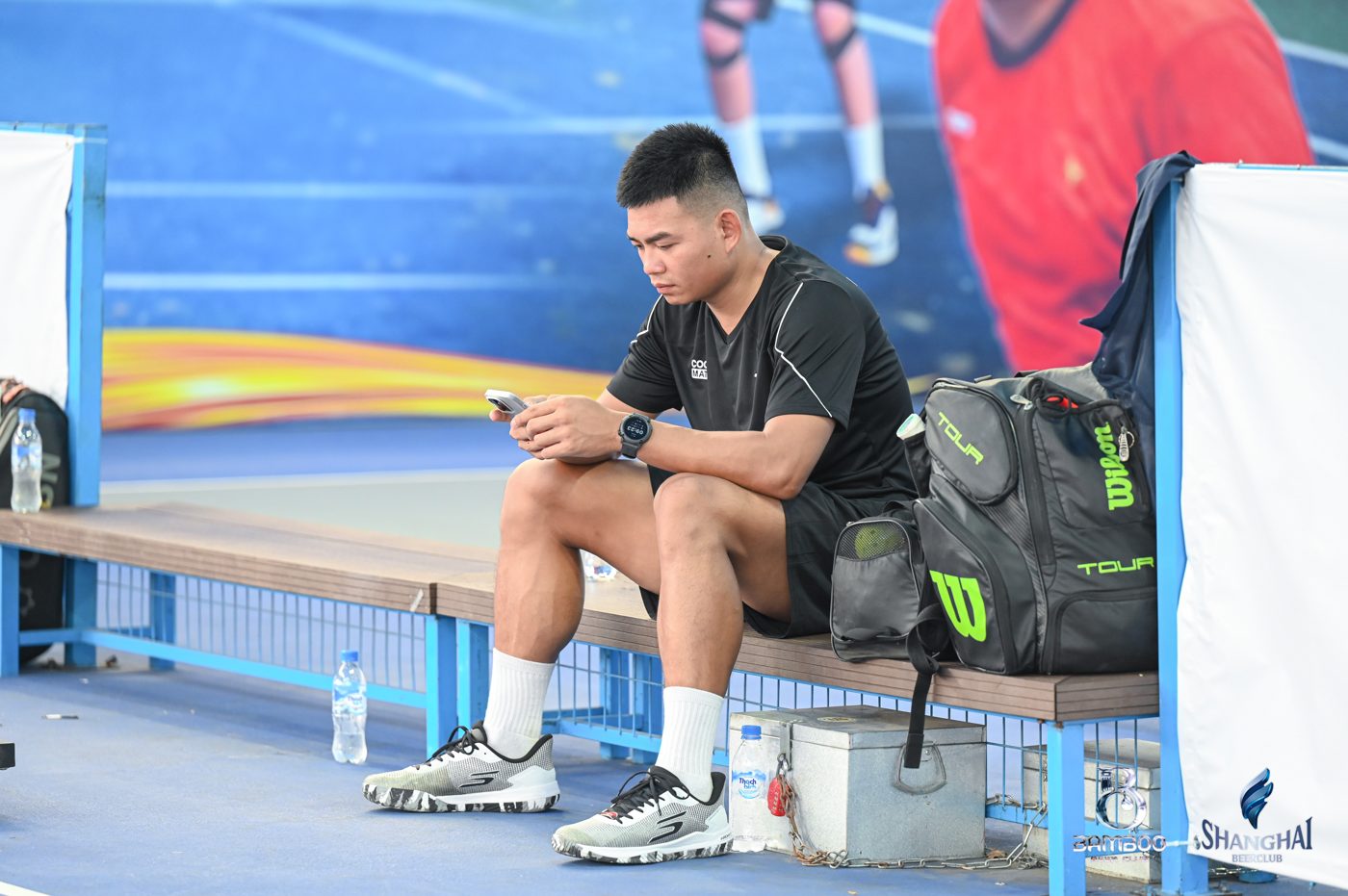 Lương Chí Bảo ngồi nghỉ bên sân, cạnh chai nước và túi vợt