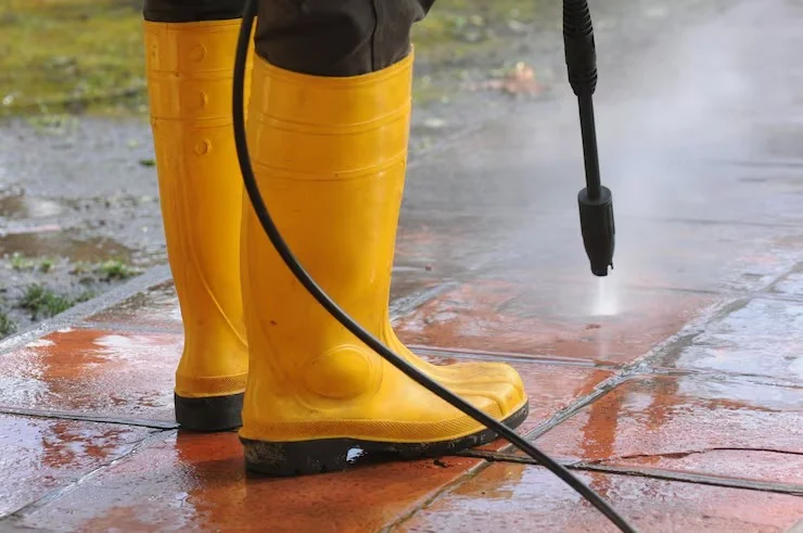 A person wearing yellow rubber boots pressure washing a dirty, wet terracotta patio with a power washer.