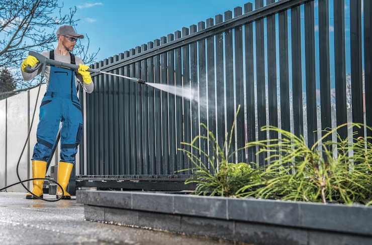 A man in blue overalls and yellow boots pressure washing a modern dark gray metal fence outdoors.