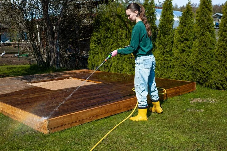 A young woman in yellow boots and jeans power washing an outdoor wooden deck surrounding a sandbox.
