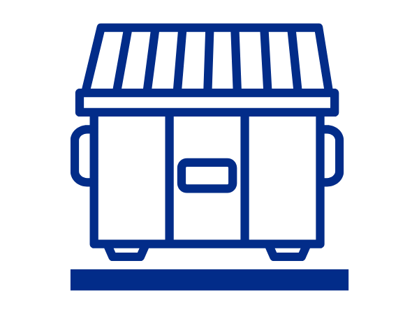 Several large black commercial waste containers are lined up along a paved sidewalk in a residential area. The bins are mounted on wheels and positioned beside a curb, with leaf-covered ground, bare trees, and apartment buildings visible in the background on an overcast day.
