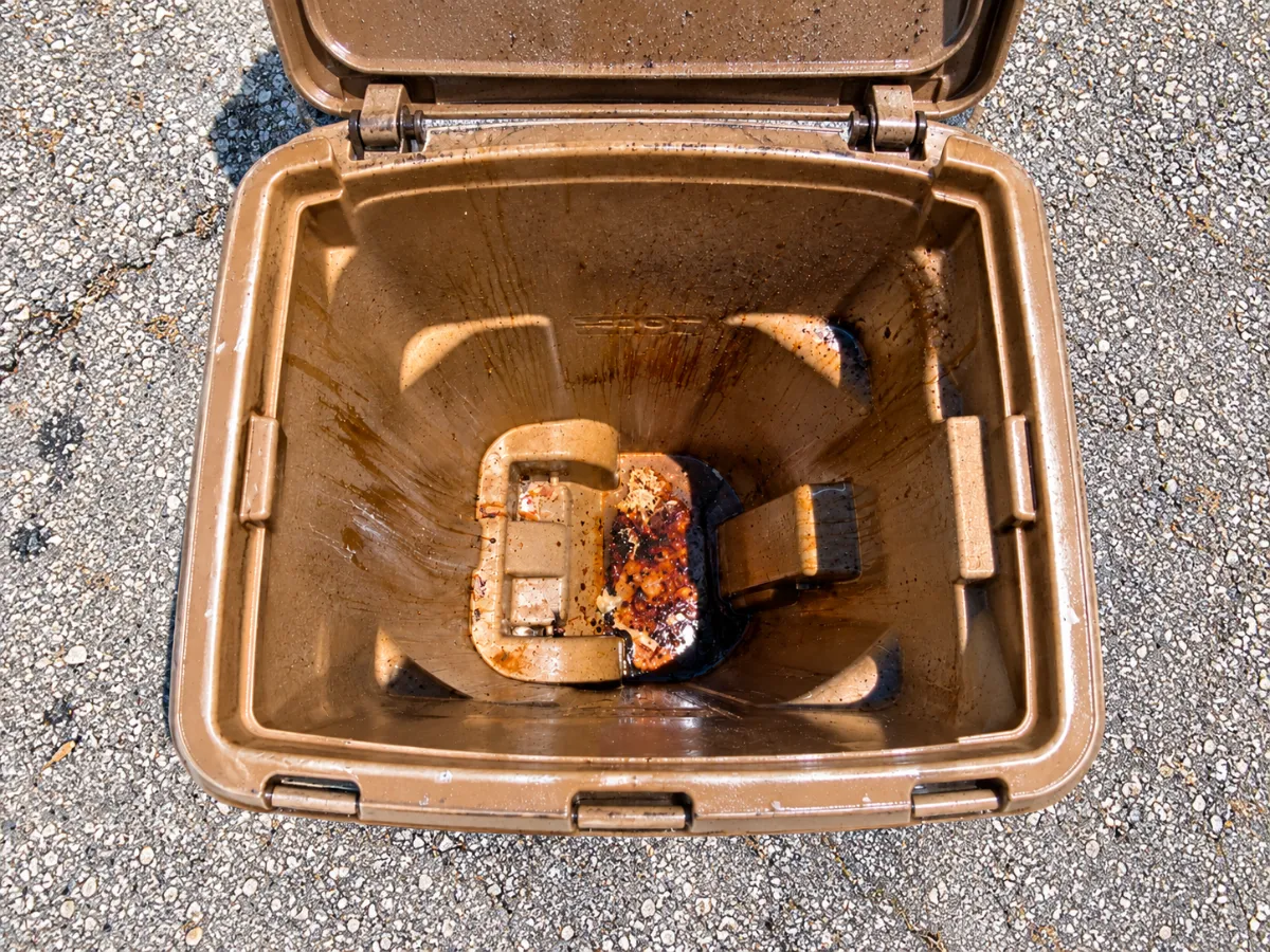 A top-down view of a dirty, open brown trash bin with grime on its interior, sitting on a gravel-like surface.
