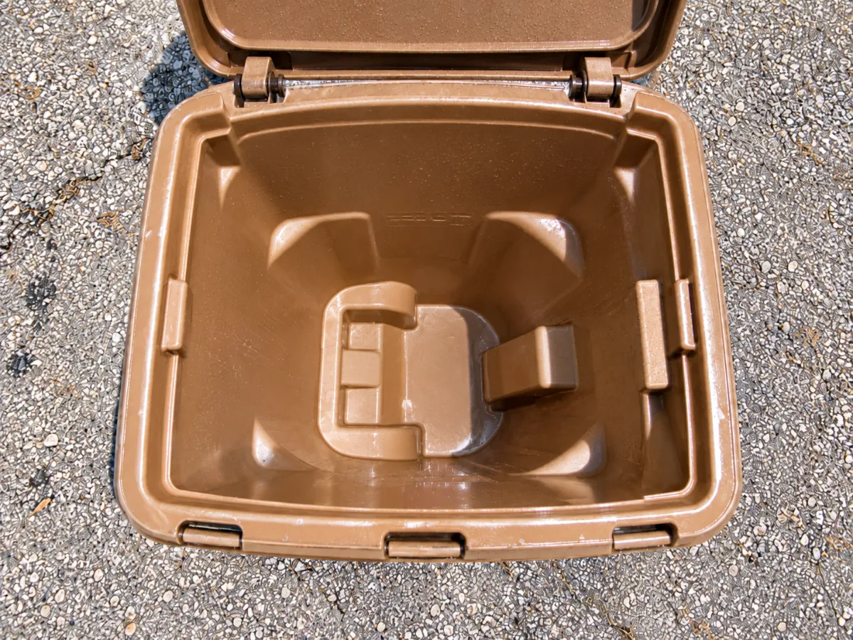 An overhead view looking down into an open, empty brown plastic bin with a hinged lid, sitting on gray gravel.