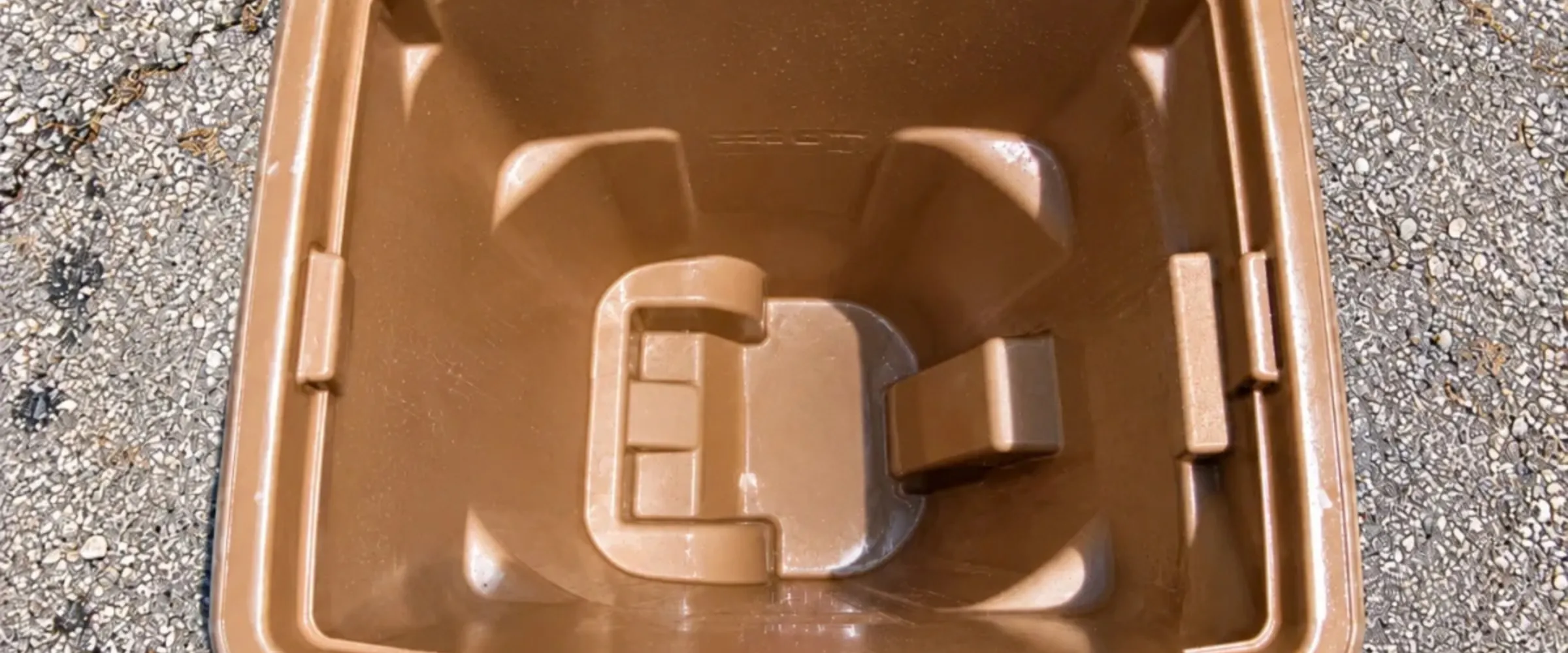 A top-down view of a rectangular brown plastic container with molded interior details, sitting on gravelly asphalt.