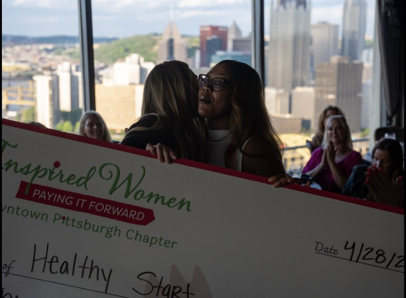 A member of Inspired Women embraces the Healthy Start representative during the ceremonial check presentation, with the Pittsburgh skyline visible through the Le Mont window