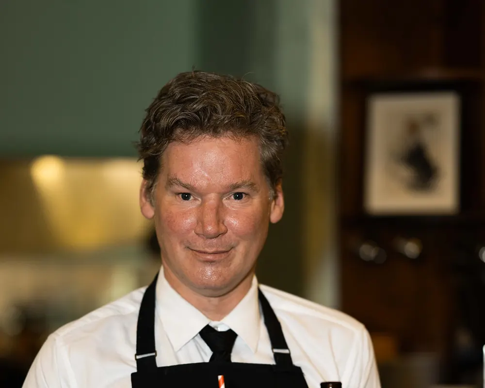 A smiling Grappino waiter in a black apron carrying a tray of drinks