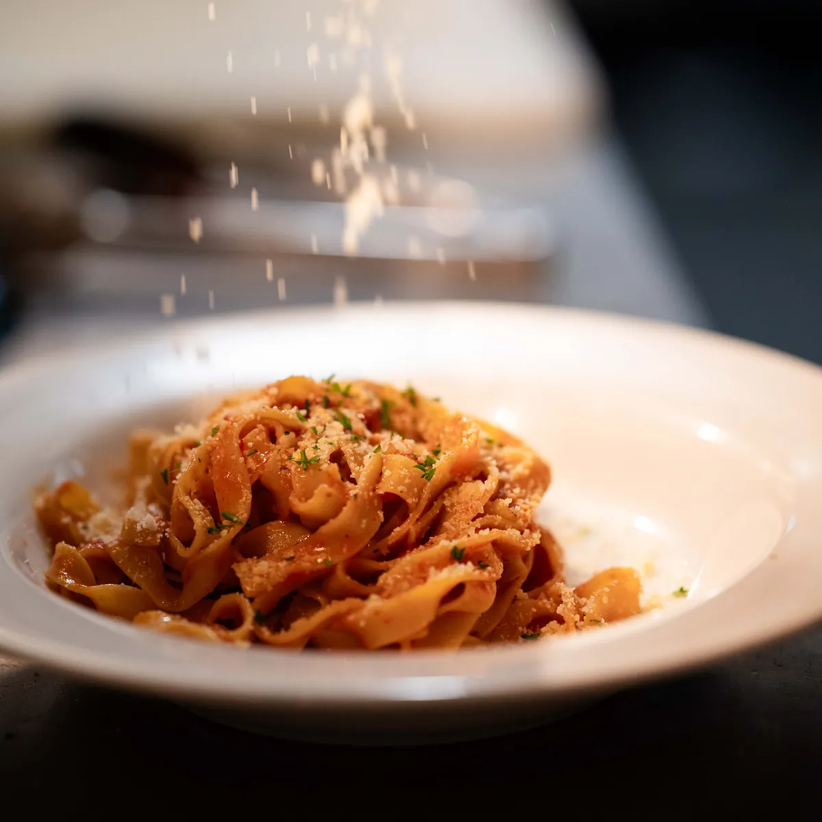 Parmesan falling over a bowl of ribbon pasta in tomato sauce at the pass in Grappino's kitchen