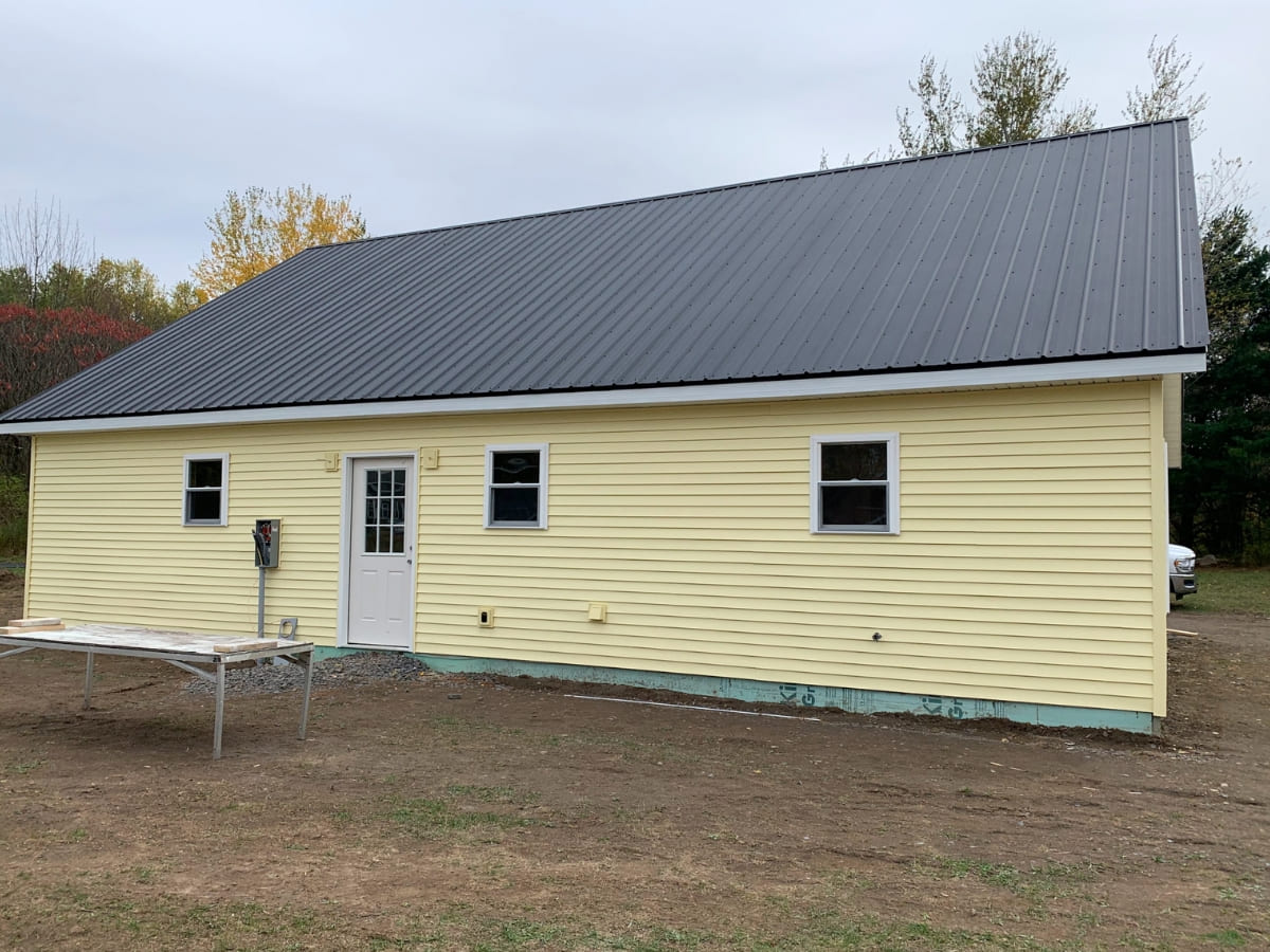 Yellow detached building with a black metal roof and white doors, set against a grassy background.