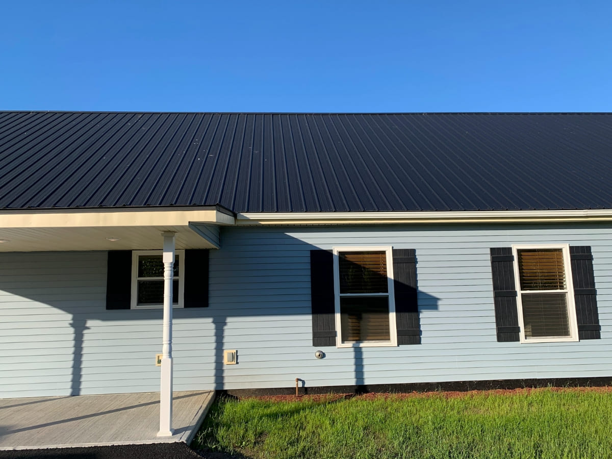 A one-story house with a dark blue metal roof, blue siding, and white trim, casting shadows in the late afternoon sun.