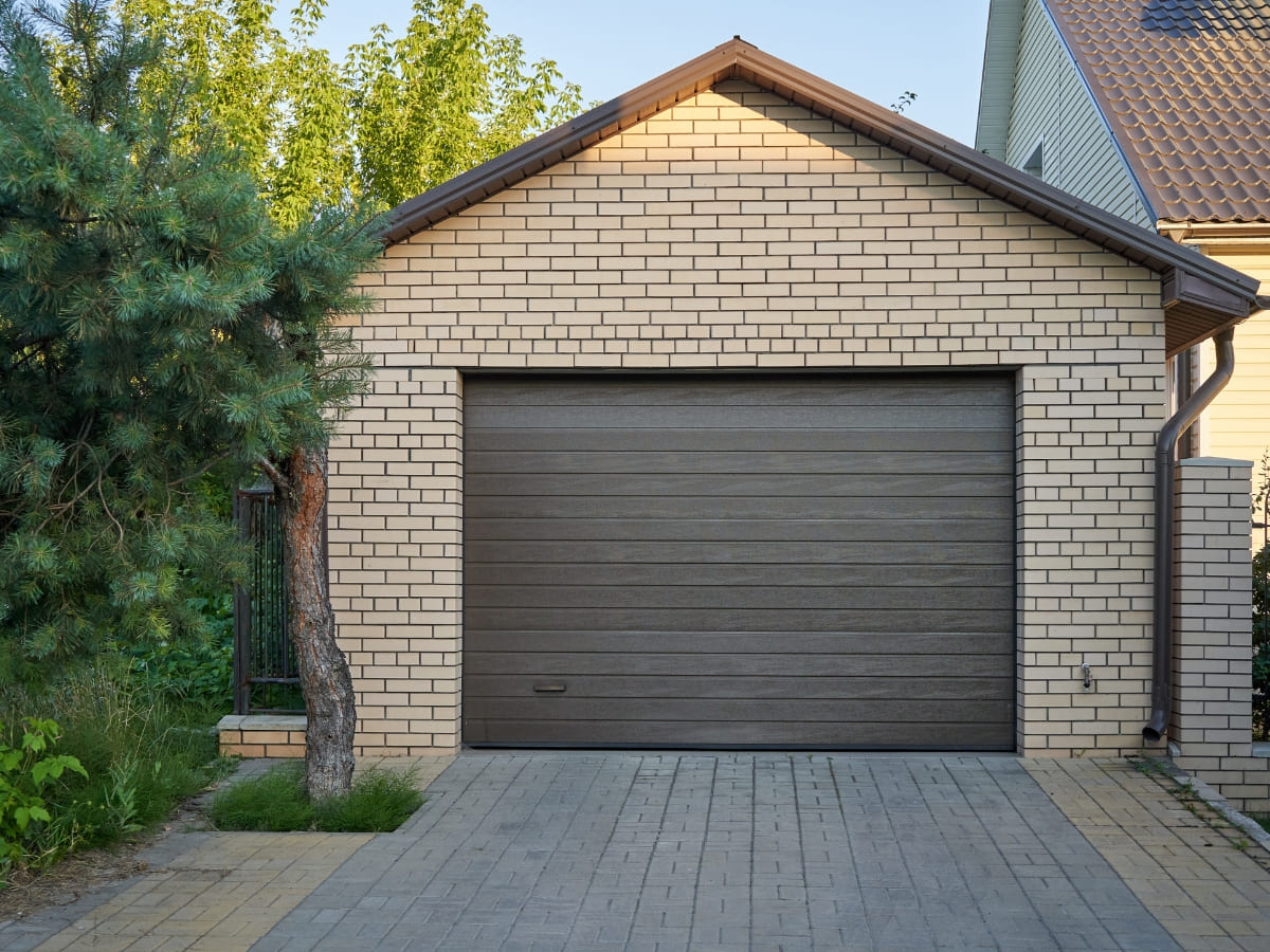 Brick garage with a brown door in a residential area, surrounded by green trees and a paved driveway.