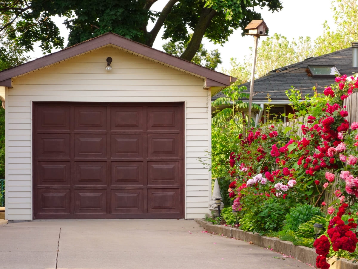 Single-car garage with a brown door and white siding, bordered by vibrant red and pink roses in a residential backyard