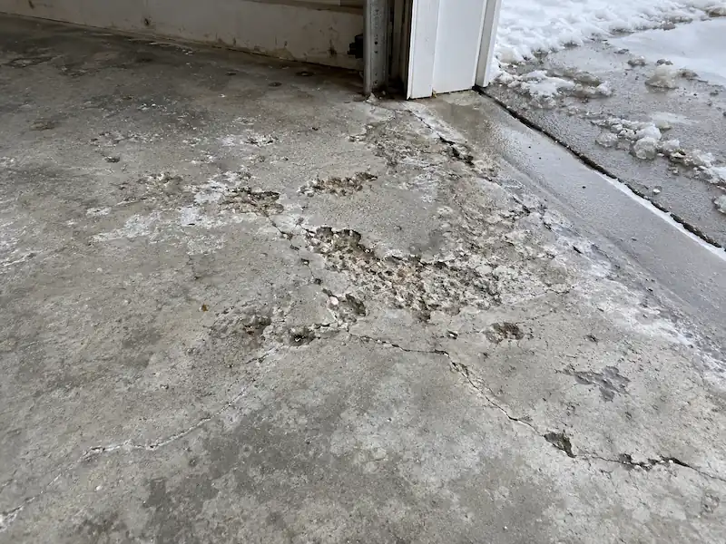 Road salt pitting, spalling and cracks in a bare concrete garage floor at the door threshold in winter