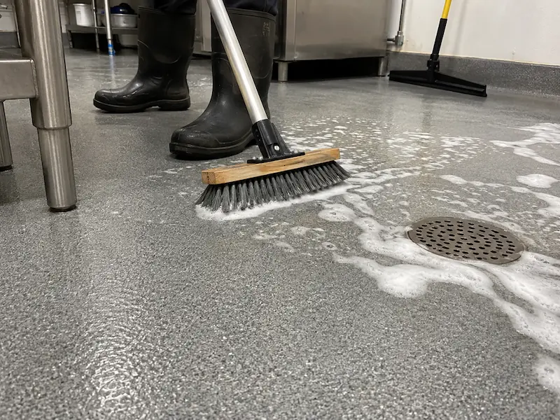 Soft-bristle deck brush scrubbing a gray quartz epoxy floor with cleaning foam beside a floor drain