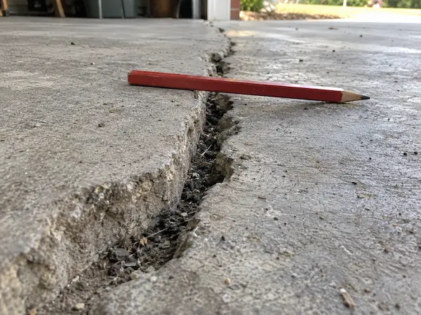 Wide crack in a concrete slab with one side sitting higher than the other, shown with a pencil laid across it