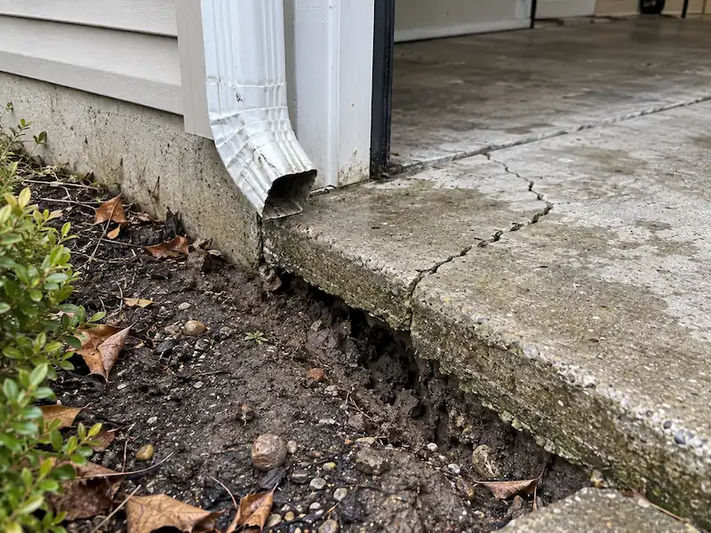 Downspout emptying at the corner of a garage slab, with soil washed out from under the cracked concrete edge
